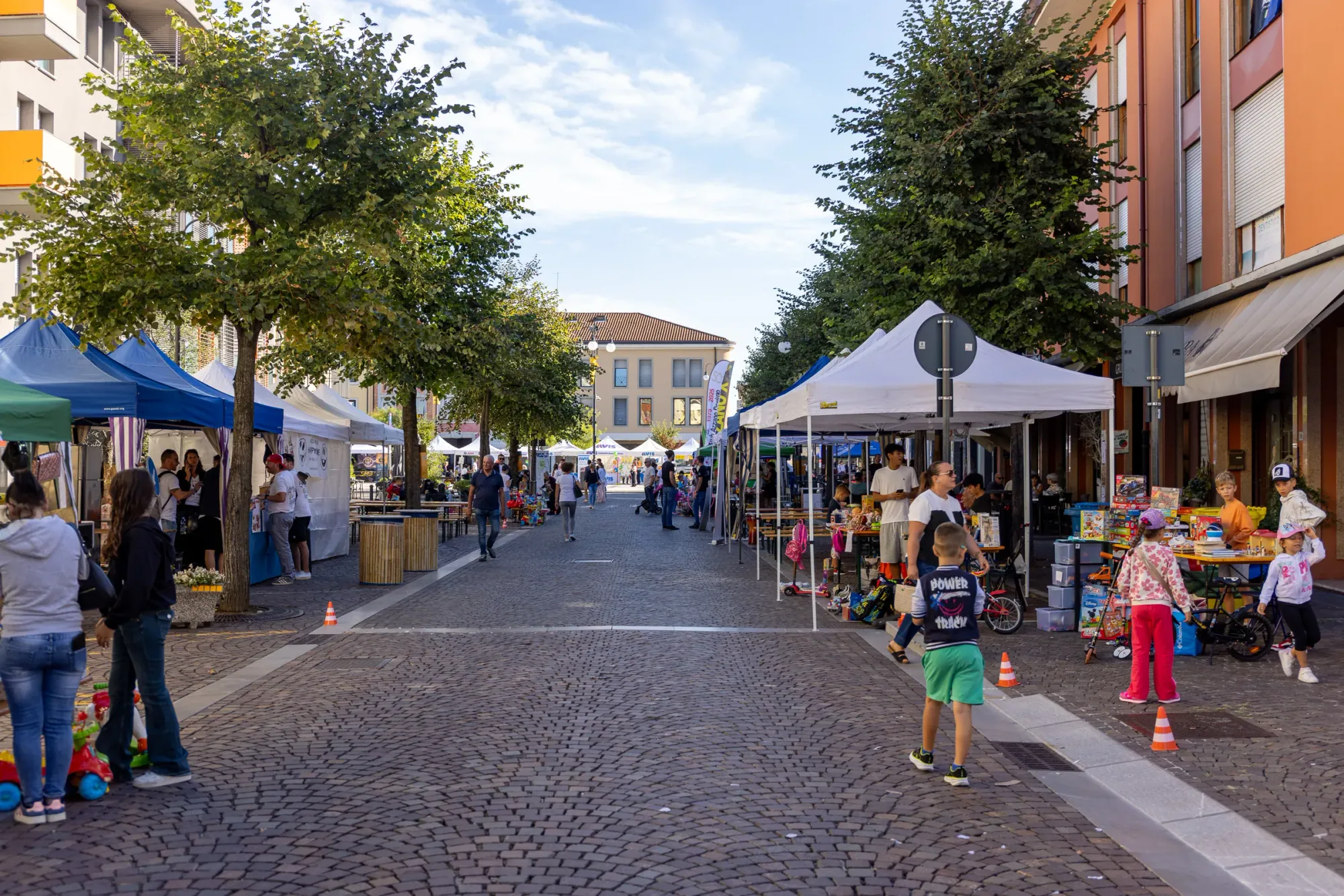 La via del mercatino vista dall'inizio: due file di gazebo e bancarelle sotto gli alberi, con le famiglie a passeggio.