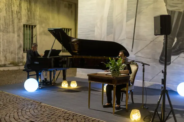 Il palco nella corte: a sinistra il pianista al pianoforte a coda, a destra la lettrice seduta a un tavolino con una pianta, davanti al microfono.