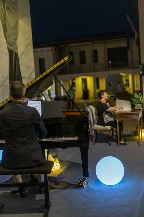 Il pianista di spalle e, oltre il pianoforte, la lettrice al tavolino; sullo sfondo i portici illuminati della corte.