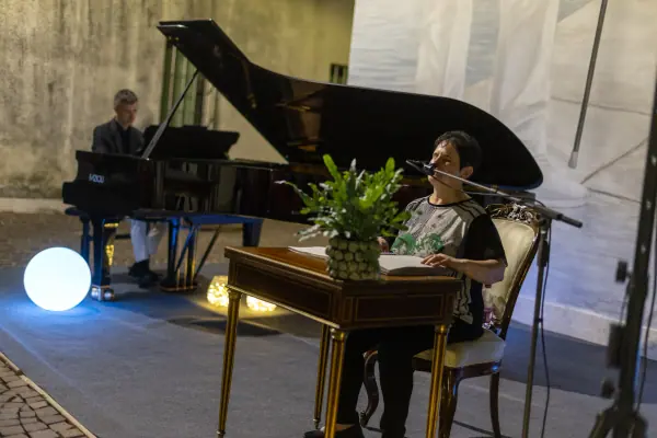 La lettrice al microfono, con le mani sul libro aperto sul tavolino; dietro di lei il pianista al pianoforte a coda.
