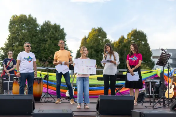 Terzo posto: sul palco, una ragazza tiene in mano il suo disegno a colori, accanto agli organizzatori e alla presentatrice al microfono.