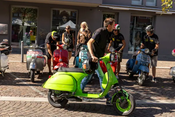 Una Vespa verde acido pronta a partire, con altri scooter d'epoca e i loro piloti con il casco in mano.