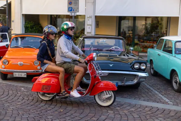 Una Vespa rossa con due passeggeri con il casco, tra le auto d'epoca in fila.