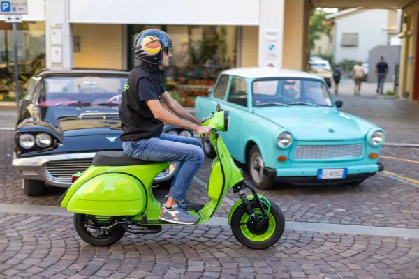 Un pilota con il casco sulla Vespa verde acido, davanti a un'auto azzurra d'epoca.