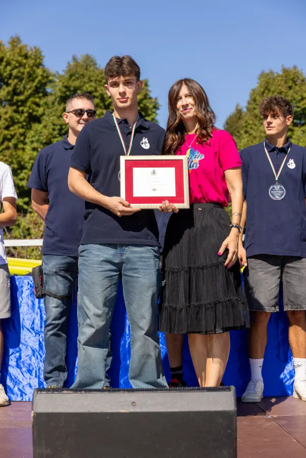 Un attestato incorniciato tenuto in mano sul palco, tra atleti con la medaglia al collo.